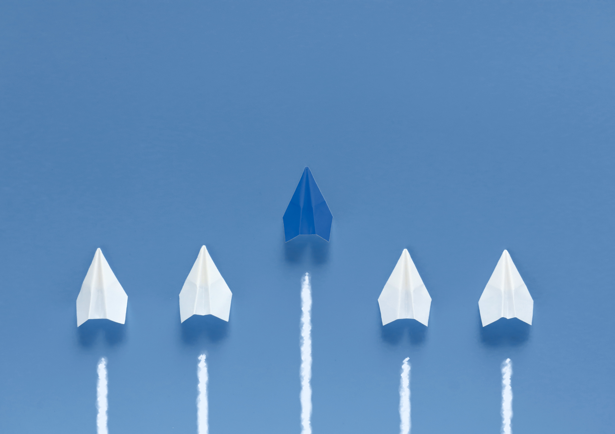 A blue paper airplane flies ahead of four white paper airplanes